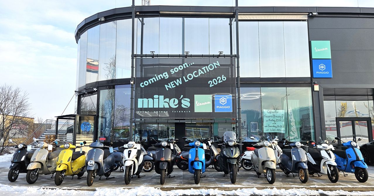 The front of a motorcycle dealership with a sign indicating 'coming soon... NEW LOCATION 2026'. Various Vespa models are parked in front.