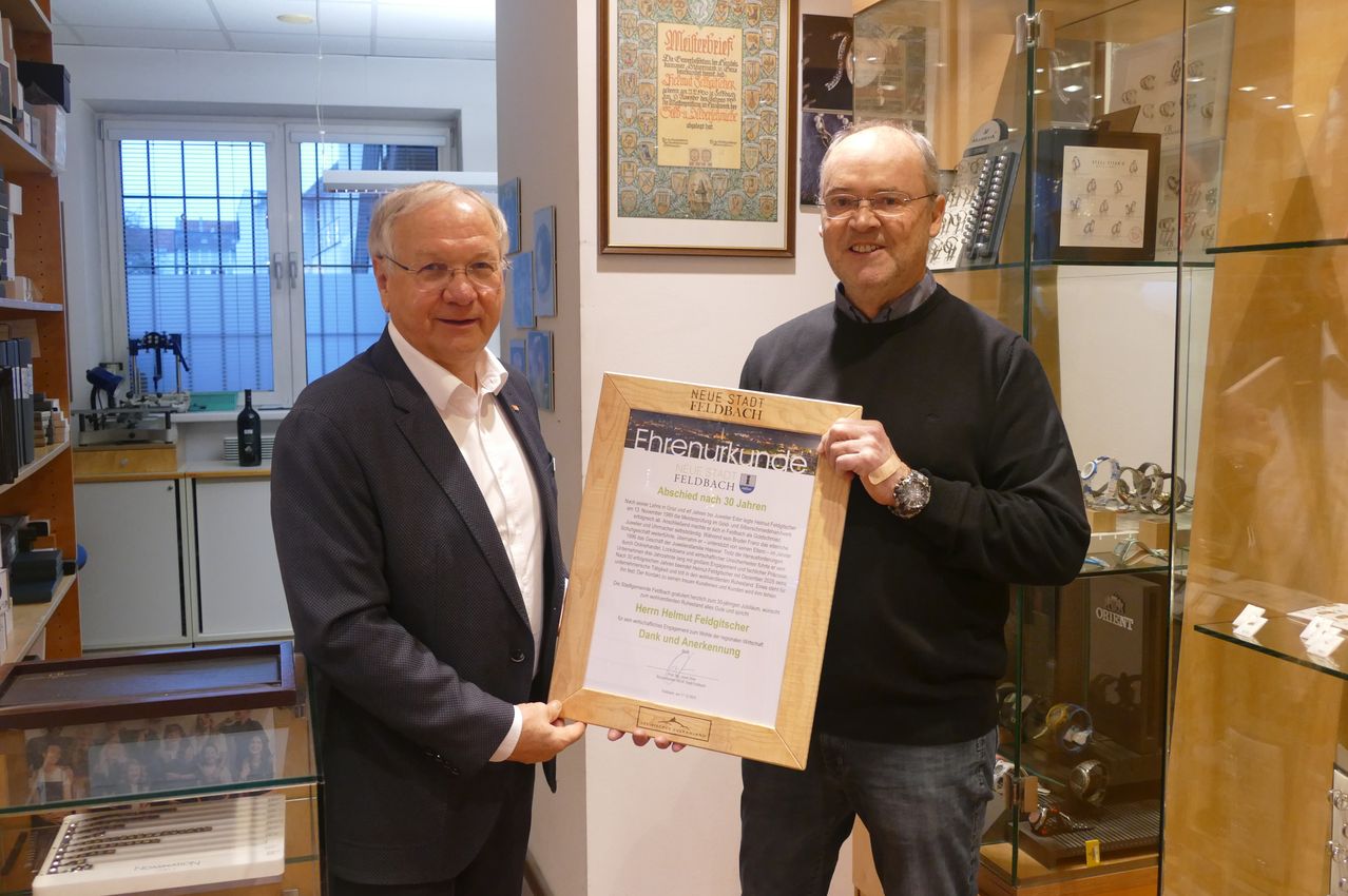 Two men stand in an office, holding a certificate. The man on the left wears a suit and a medal, while the man on the right wears glasses and a watch. Behind them, a glass cabinet displays various items.