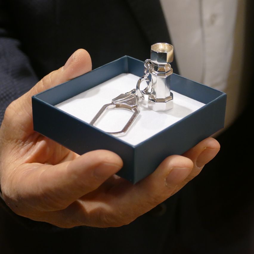 A person is holding a blue box with a silver keychain inside. The keychain has a cylindrical shape with a flat top and a small chain. The box has a white interior.