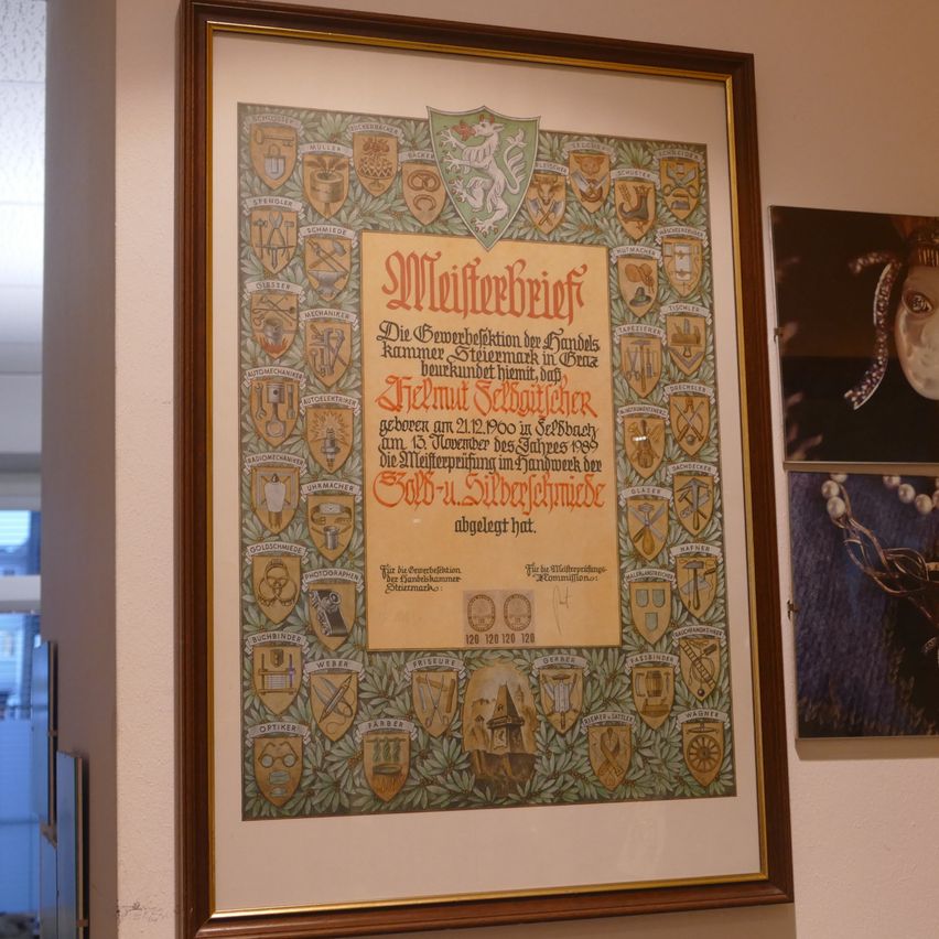 A framed certificate hangs on a wall, featuring a golden crest and various emblems. It is titled 'Master Certificate' and lists a name, date, and a sign of approval. Two additional framed pictures are also on the wall.