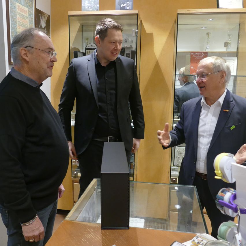 Three men are in a room with glass cabinets. The man in the middle is gesturing while speaking. The man on the left is wearing glasses and a black sweater. The man on the right is wearing a suit and tie.