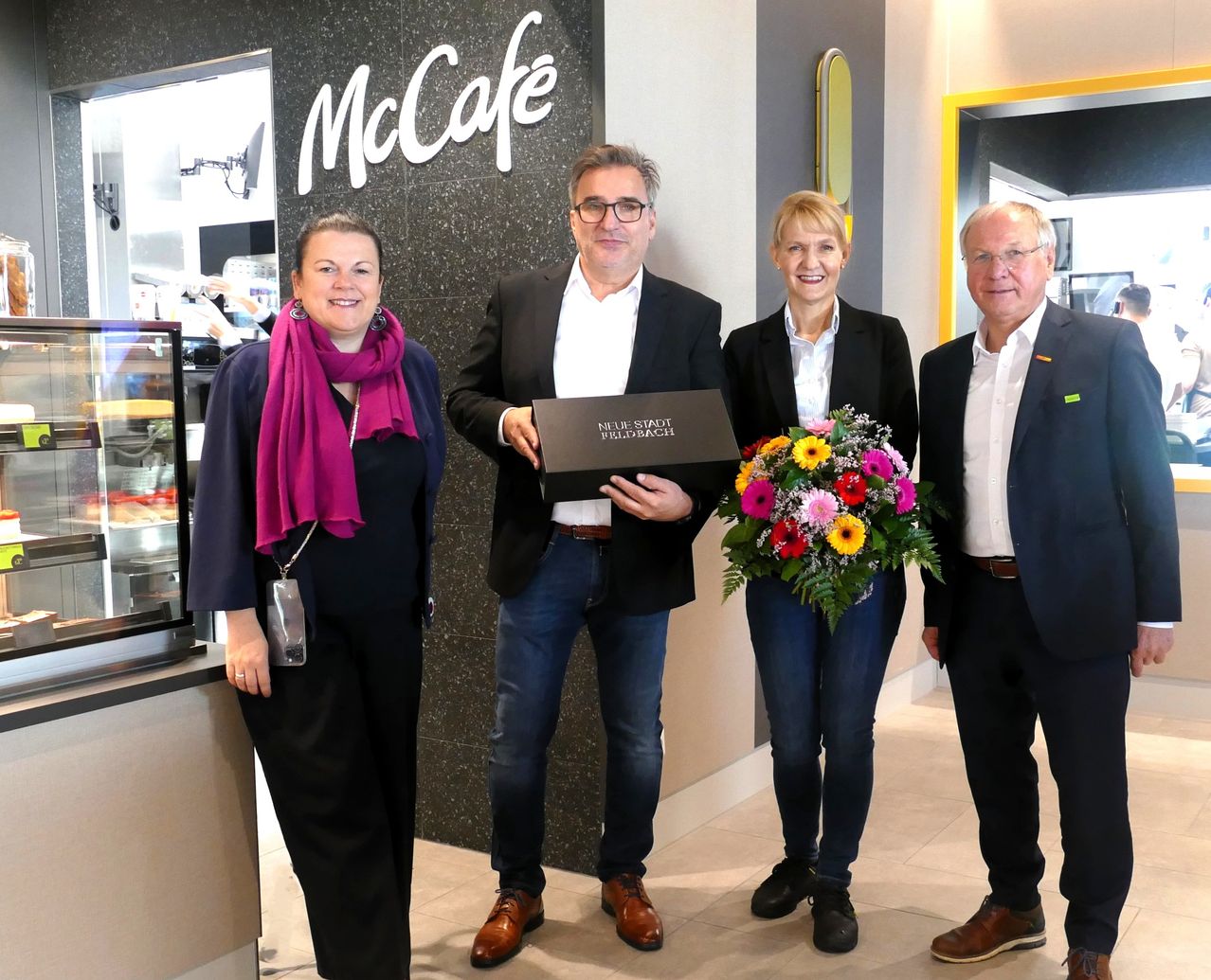 Four people stand in front of a McCafe, smiling. The man in the middle holds a black box. A woman on the right holds a bouquet of flowers.