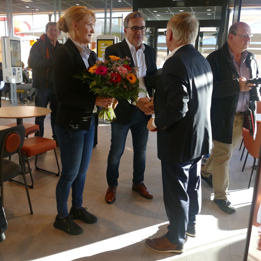 Five people are standing inside a room. One woman is holding a bouquet of flowers. Another man is holding a pen and paper, and he is shaking hands with the woman.
