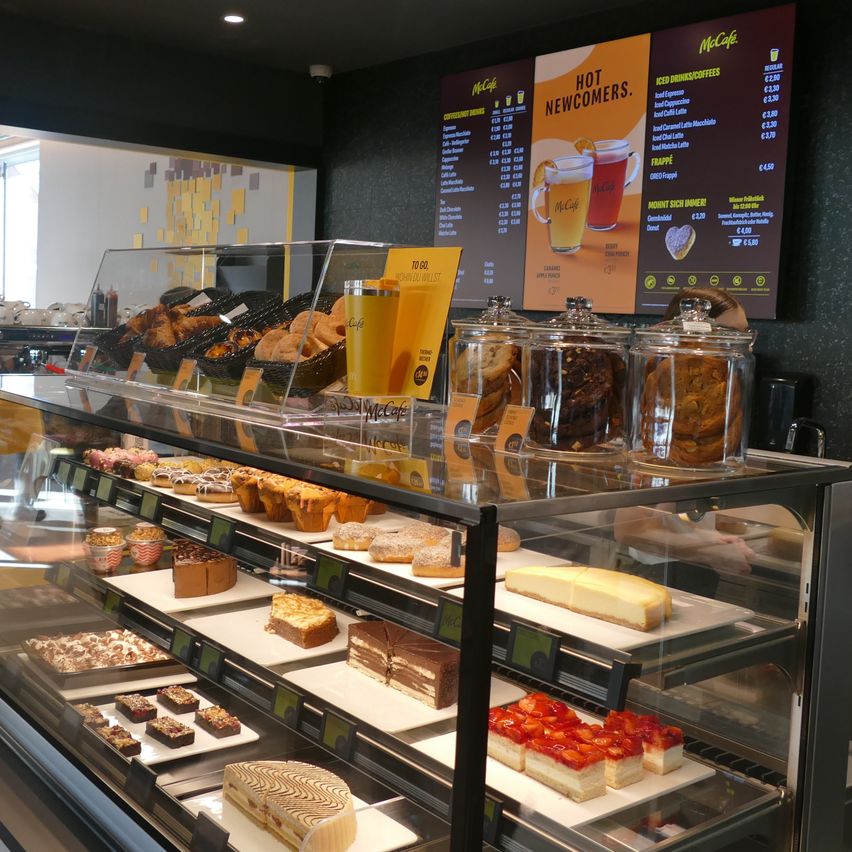 A bakery section with pastries and desserts on display in a glass case. A menu board behind the display offers hot newcomers and iced drinks.