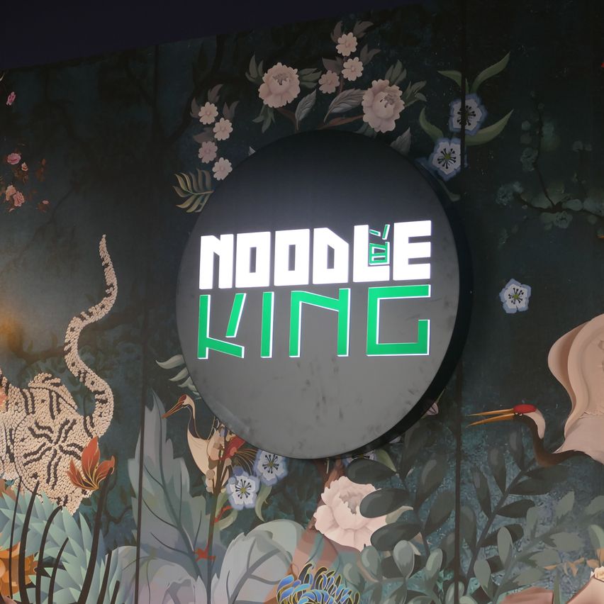 Ein rundes Schild mit den Worten Noodle King in Grün und Weiß auf schwarzem Hintergrund, umgeben von einem Blumendesign mit rosa und weißen Blüten.