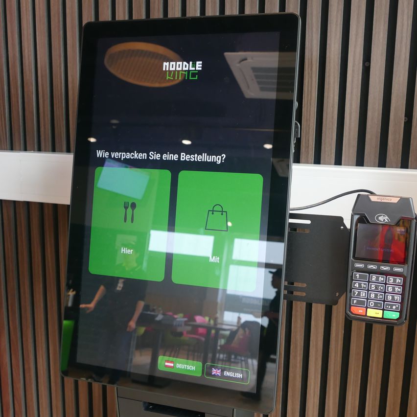 Ein Touchscreen-Interface von Noodle King zeigt Optionen zum Bestellen von Speisen zum Essen vor Ort oder zum Mitnehmen. Der Bildschirm hat Optionen für die Sprachen Deutsch und Englisch.