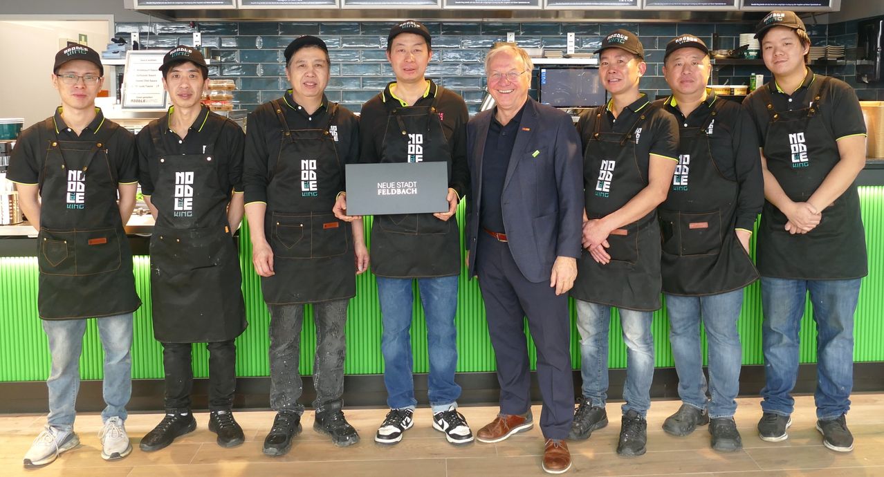 Fünf Männer, gekleidet in schwarzen Schürzen, posieren für ein Foto mit einem lächelnden Mann in einem Anzug. Sie stehen vor einer grünen Wand, und der Mann im Anzug hält eine Plakette, auf der 'Neue Stadt Feldbach' steht.
