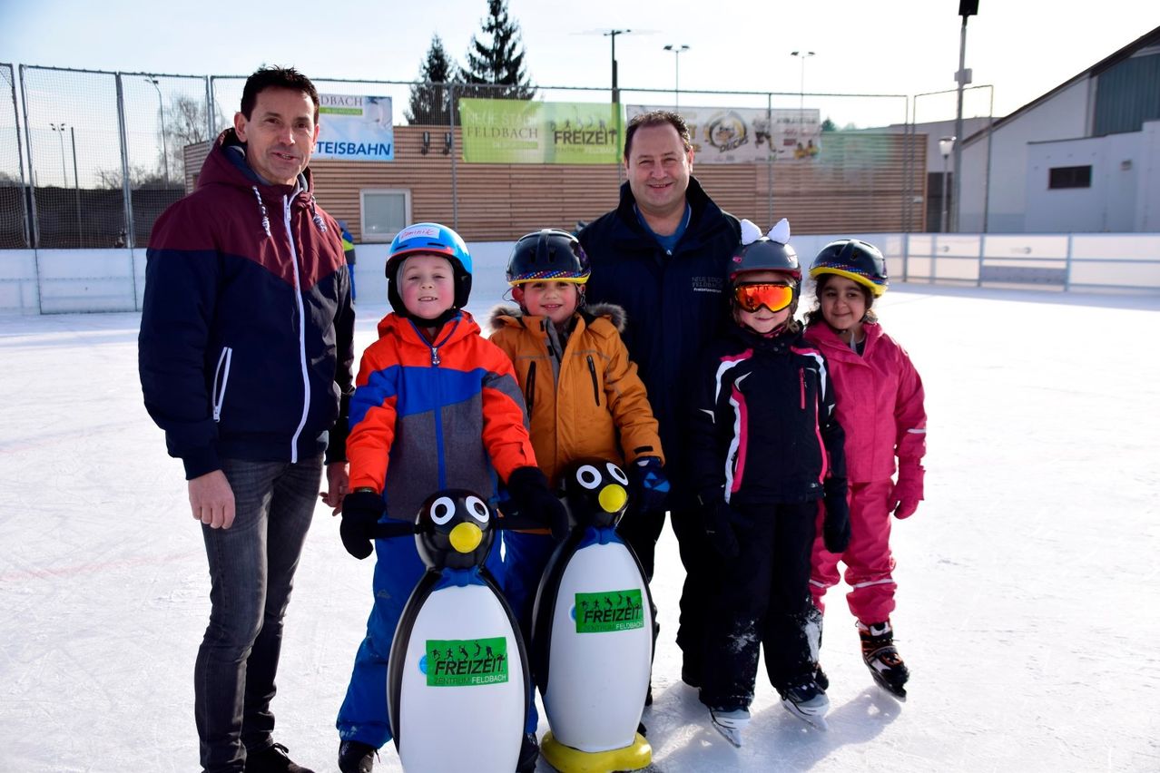 Vier Erwachsene und vier Kinder stehen auf einer Eisbahn. Die Kinder tragen Helme und halten pinguinförmige Objekte. Sie sind in Winterkleidung gekleidet. Hinter ihnen steht ein Zaun mit Bannern.