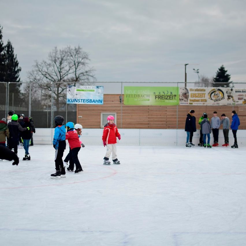 Mehrere Menschen skaten auf einer Eisbahn im Freien. Einige tragen Helme und Jacken. Im Hintergrund sind ein Zaun und Plakate zu sehen.