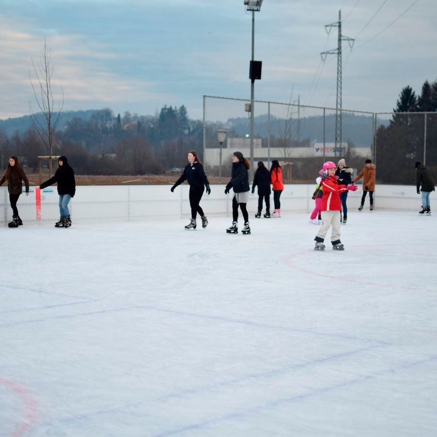 Eine Gruppe von Menschen eislaufen auf einer Freiluftbahn. Einige tragen Jacken und Handschuhe, und es gibt Zäune und Bäume im Hintergrund.