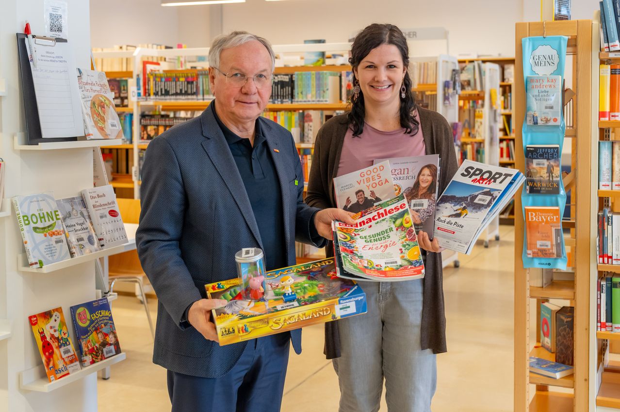 Ein älterer Mann und eine Frau stehen in einer Bibliothek, halten Zeitschriften und ein Brettspiel in den Händen. Der Mann trägt einen dunklen Anzug und hält einen kleinen Behälter. Die Frau lächelt warm und trägt ein helles Oberteil. Hinter ihnen sind mit Büchern gefüllte Regale zu sehen.
