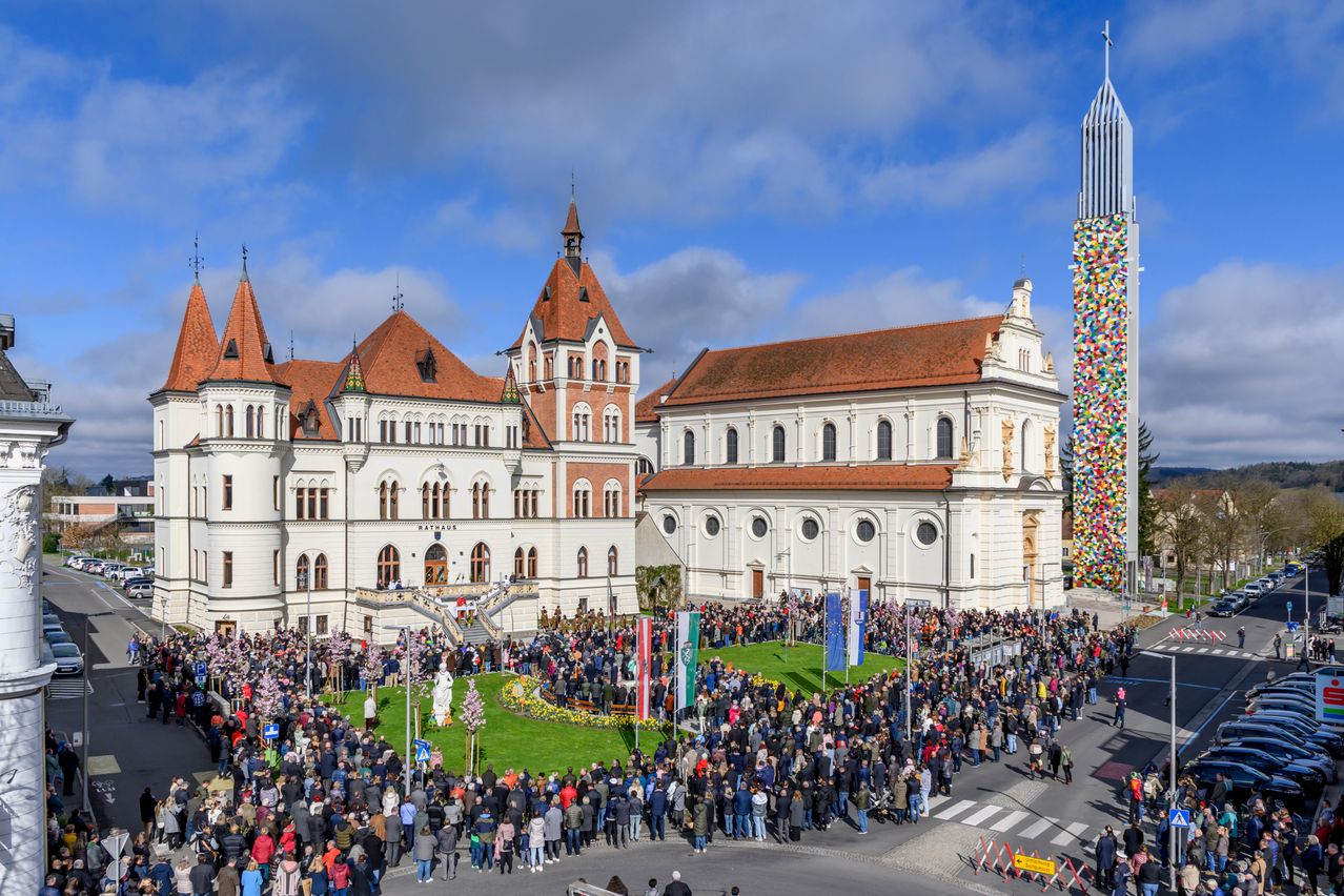 Eine große Menschenmenge versammelt sich vor einem historischen Gebäude mit roten Dächern und aufwendigen Details, Flaggen und einer Statue in der Mitte unter blauem Himmel.