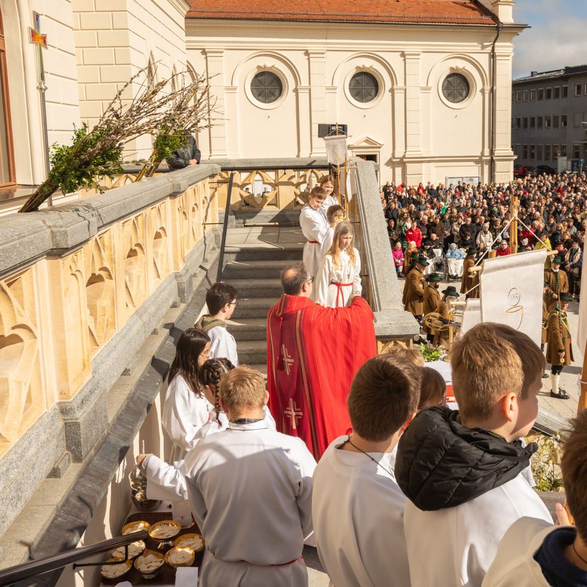 Eine religiöse Zeremonie vor einer großen Menschenmenge vor einer Kirche. Teilnehmer in weißen Gewändern und ein Priester in Rot. Stufen führen zum Altar mit einem Banner.