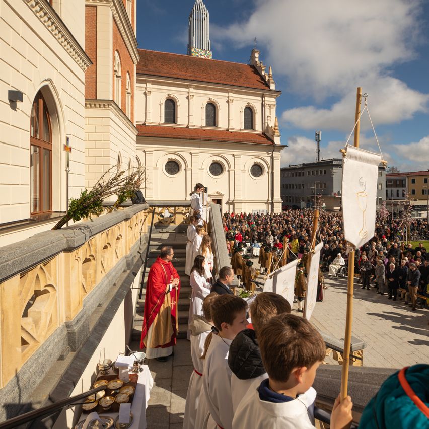 Eine religiöse Prozession vor einer Kirche, mit einem Priester in roten Roben und Messdienern in Weiß. Eine große Menge versammelt sich, einige halten Banner, unter blauem Himmel.