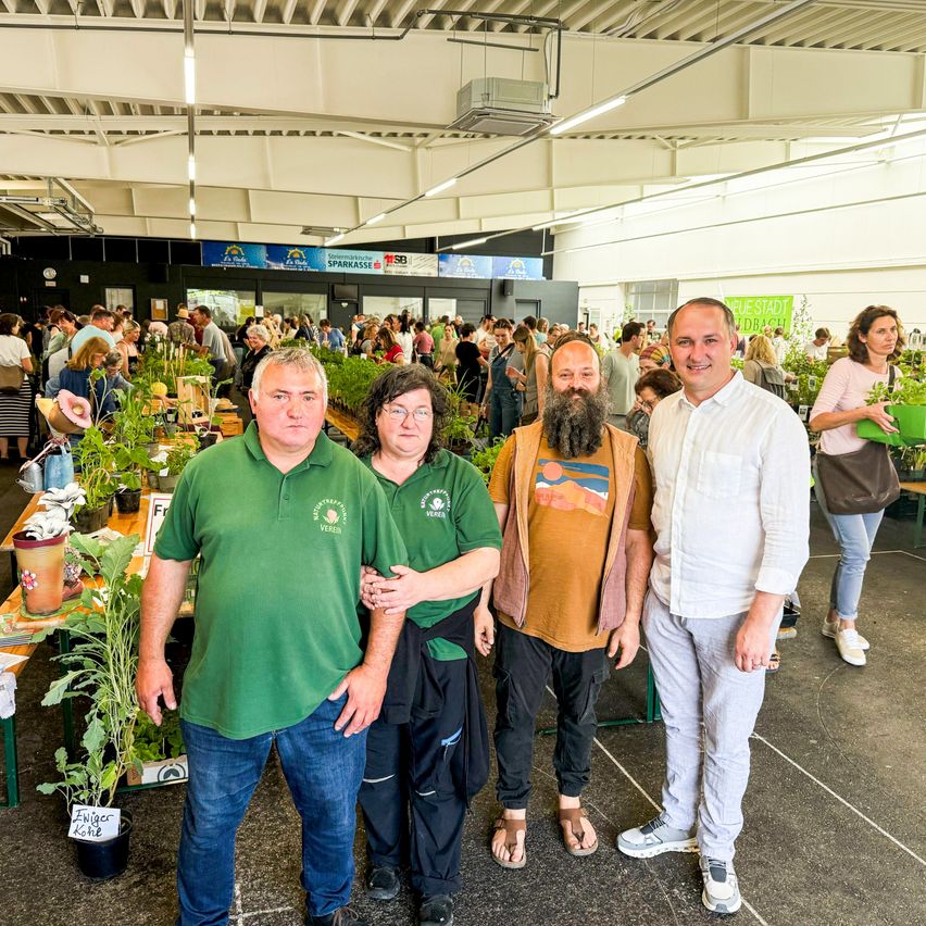 Vier Personen posieren für ein Foto in einem Raum mit vielen Topfpflanzen und einkaufenden Menschen. Zwei von ihnen tragen grüne Hemden, und einer hat einen Bart.
