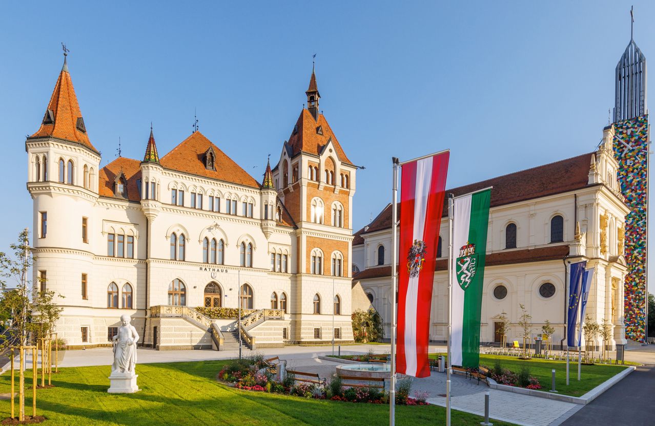 Ein historisches Gebäude mit einer Statue davor und Fahnen an Stangen. Es hat einen Turm, viele Fenster und ist von grünem Rasen und Blumen umgeben.