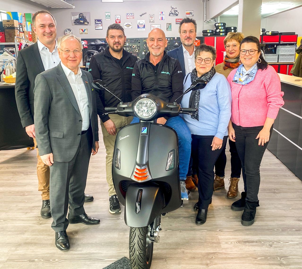 Eine Gruppe von Menschen posiert für ein Foto mit einem Roller in einem Geschäft. Sie tragen formelle und lässige Kleidung.