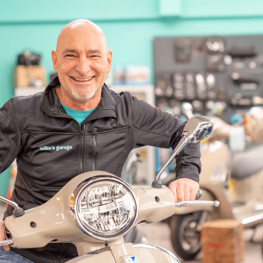 Ein kahlköpfiger Mann in einem schwarzen Jackett sitzt auf einem hellen Roller und lächelt. Hinter ihm sind eine türkisfarbene Wand und verschiedene Roller und Gegenstände zu sehen.