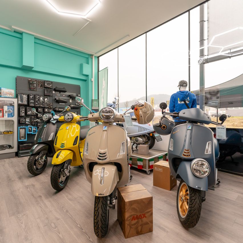 Ein Ausstellungsraum mit mehreren Scootern, die in verschiedenen Farben geparkt sind. Ein blauer Kapuzenpullover befindet sich in der Nähe. Zubehör und Helme sind an der Wand ausgestellt.