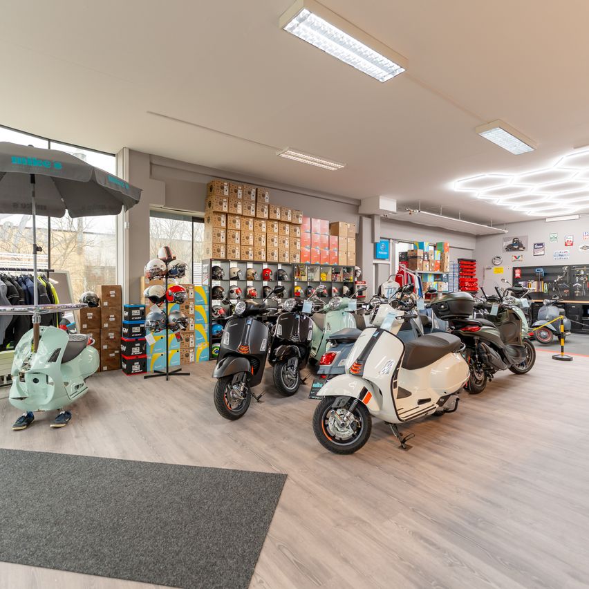 Ein Showroom mit mehreren Rollern, Helmen und Jacken zur Schau gestellt. Der Boden ist aus Holz, und ein großer Regenschirm ist sichtbar.