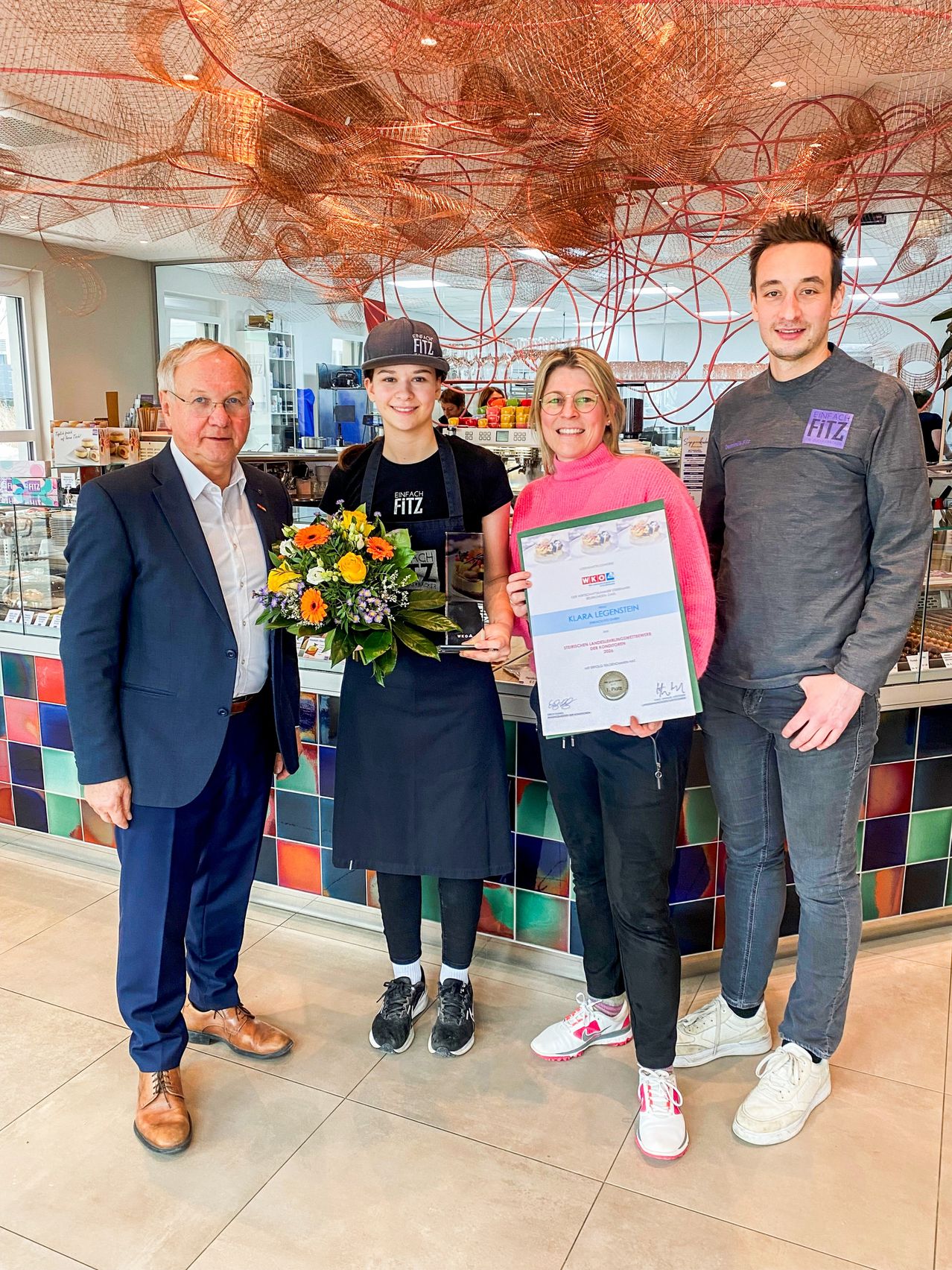 Vier Menschen stehen vor einer Theke. Eine Frau hält ein Zertifikat und Blumen. Ein Mann neben ihr trägt ein Schürzen. Der Mann rechts trägt ein graues Shirt mit FITZ darauf.