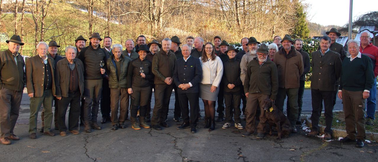 Eine Gruppe von Menschen in Winterkleidung, einschließlich Jacken und Mützen, posiert für ein Foto in einem bewaldeten Gebiet. Eine Frau hält ein Weinglas.