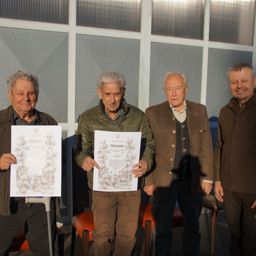 Vier ältere Männer stehen vor einer Glaswand und halten Urkunden in den Händen. Der Mann auf der linken Seite hält eine Urkunde mit einem Hirschdesign. Sie alle lächeln.