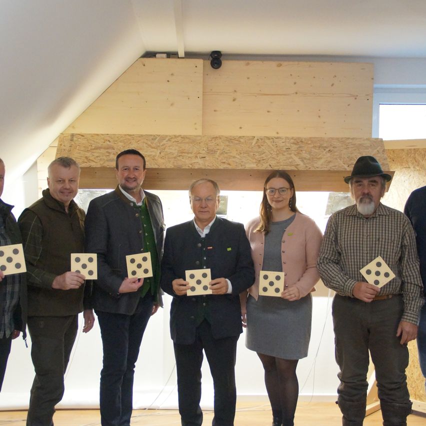 Sechs Menschen stehen in einer Reihe und halten Würfel. Hinter ihnen befindet sich eine Holzkonstruktion, und eine Person trägt einen Hut.