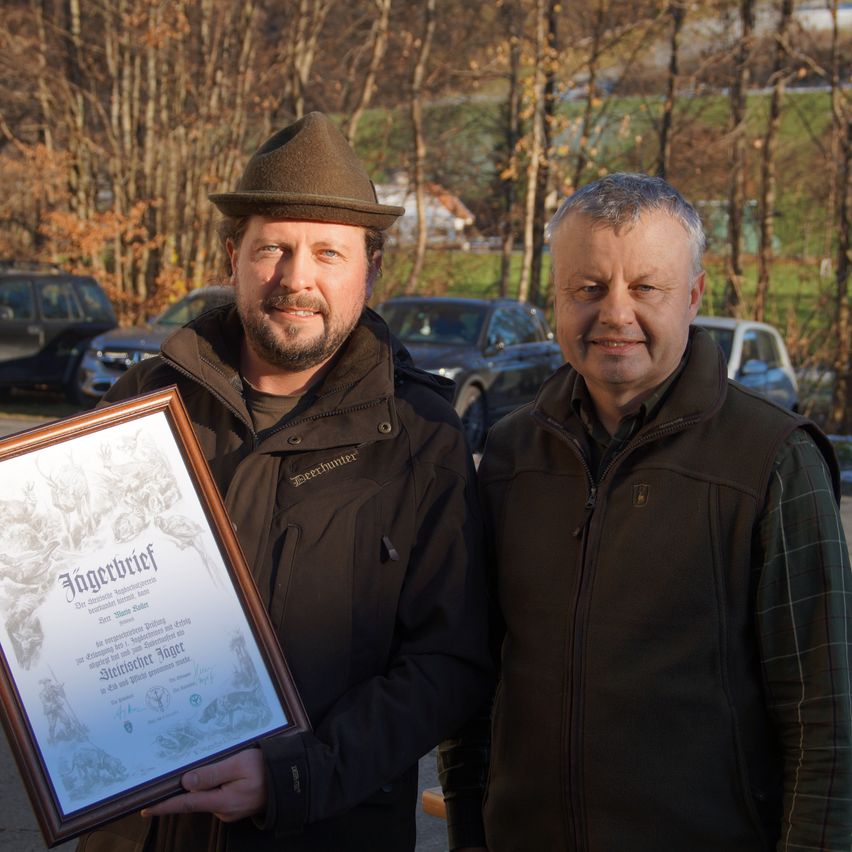 Zwei Männer stehen draußen in einem Park. Der Mann auf der linken Seite hält ein gerahmtes Zertifikat. Der Mann auf der rechten Seite trägt eine Jacke. Hinter ihnen sind Bäume, Gras und geparkte Autos zu sehen.