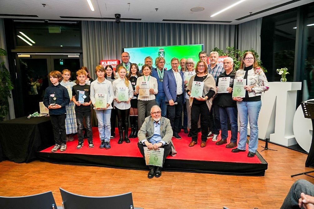 Eine Gruppe von Menschen steht auf einer Bühne und hält Urkunden. Ein älterer Mann sitzt vorne, und ein großer Bildschirm ist hinter ihnen.