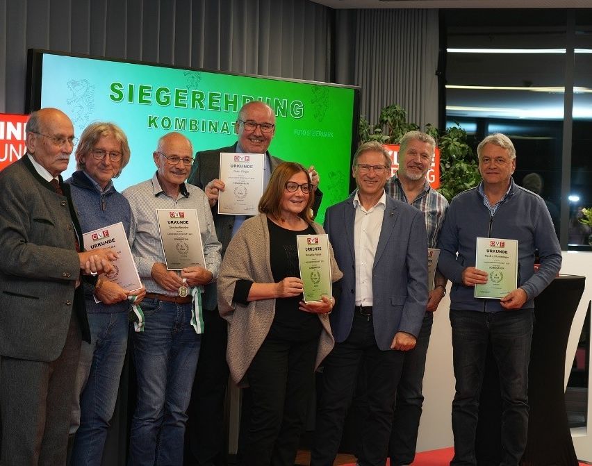 Acht Menschen posieren für ein Foto mit Urkunden. Hinter ihnen steht ein grüner Banner mit der Aufschrift 'SIEGEREHRUNG KOMBINAT'. Zwei Männer in der Mitte halten Urkunden mit Logo und Text.
