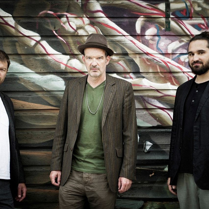 Three men stand together in front of a graffiti-covered wooden wall. The man in the middle wears a hat and necklace, while the others wear jackets.