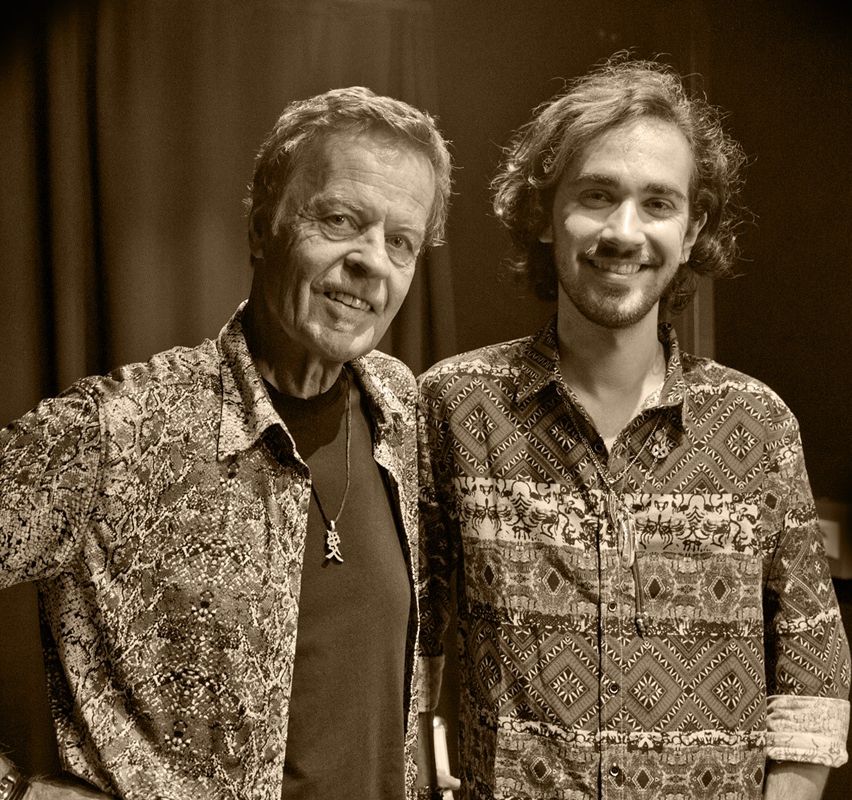 Two men, one older and one younger, stand side by side, smiling for a photo. The older man wears a patterned shirt and a necklace. The younger man has long curly hair and also wears a patterned shirt. They are in front of a dark background.