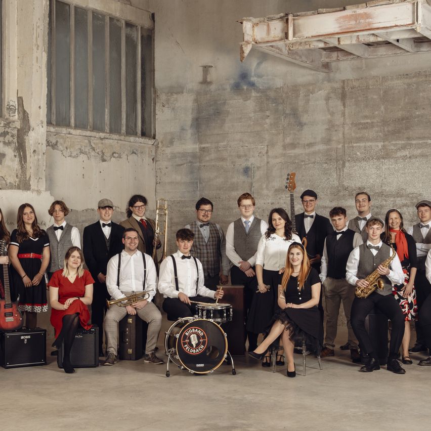 A group of musicians is standing and sitting inside a room with concrete walls. They are holding musical instruments and wearing formal attire. The man sitting on the drum is looking at the camera.