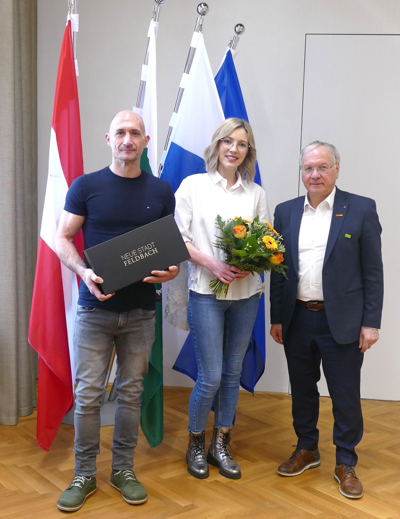 Drei Personen stehen vor Flaggen; ein Mann hält eine Box, eine Frau Blumen und ein weiterer Mann steht neben ihnen.