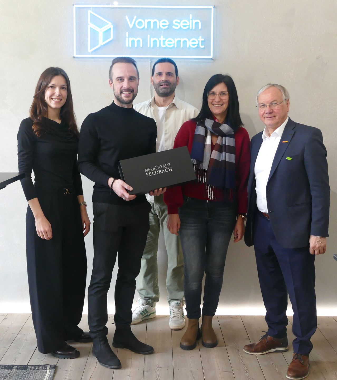 Fünf Menschen stehen lächelnd zusammen und halten eine Box. Hinter ihnen ist eine weiße Wand mit einem Schild, das 'vorne sein im Internet' sagt. Die Person in der Mitte hält die Box mit den Worten 'Neue Stadt Feldbach' darauf.