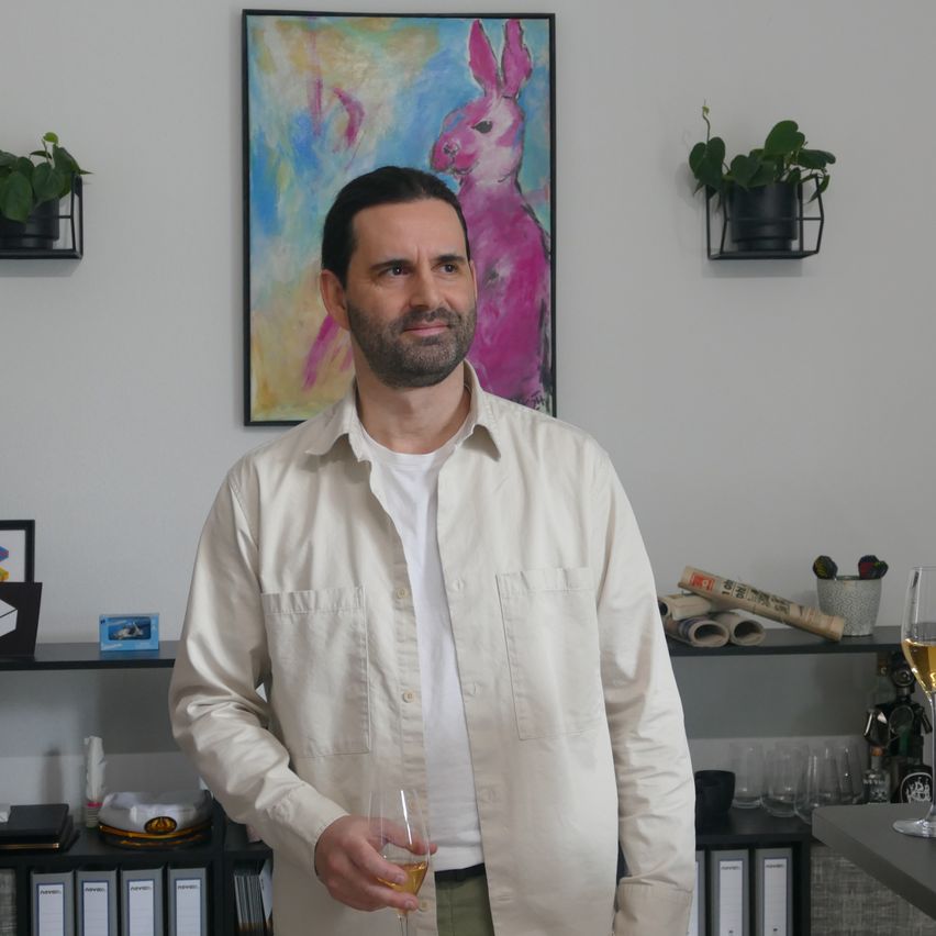 Ein Mann steht drinnen, lächelt und hält ein Weinglas. Hinter ihm zeigt eine Wand ein rosafarbenes Hasenbild und Topfpflanzen. Unten befindet sich ein Regal mit verschiedenen Gegenständen.