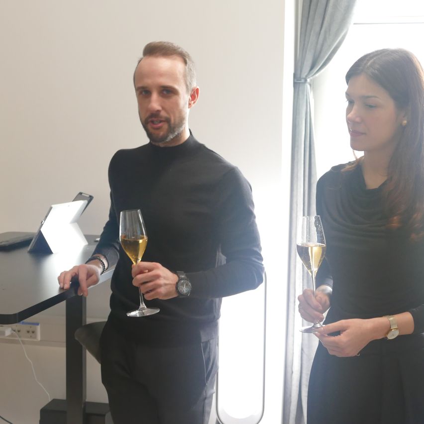 Ein Mann und eine Frau stehen in einem Büroraum und halten Weingläser in der Hand. Der Mann trägt eine Uhr. Hinter ihnen steht ein Schreibtisch mit Laptop und Steckdose.