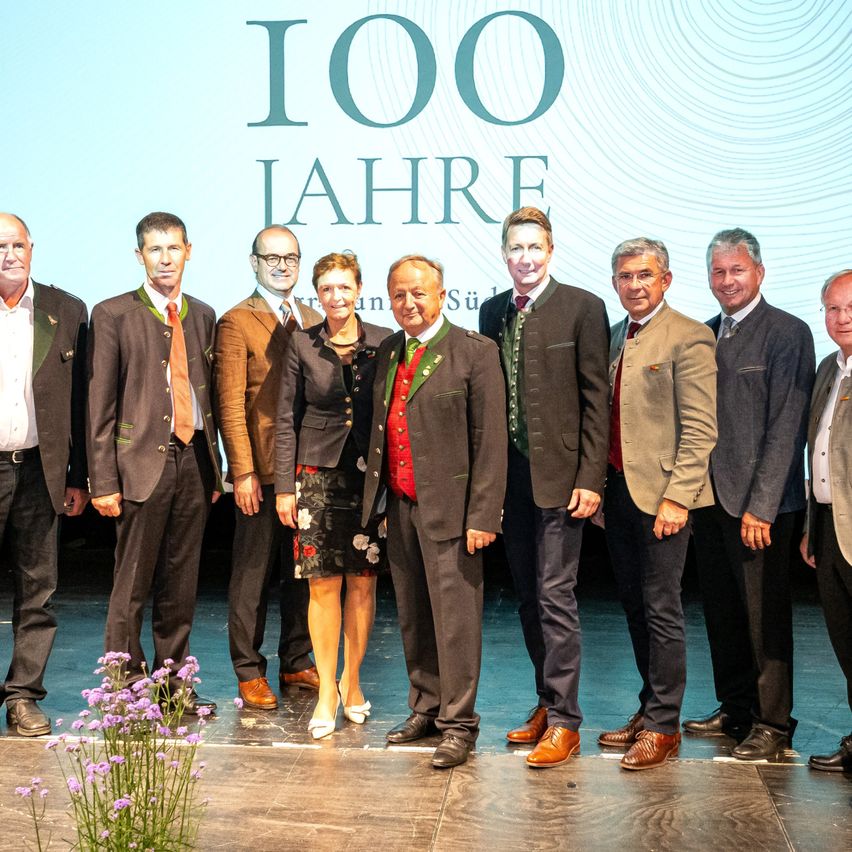 A group of formally dressed individuals stand on a stage, with a large backdrop displaying the text '100 Jahre'.