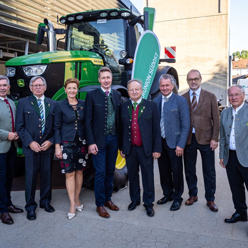 A group of formally dressed individuals stand in front of a large green tractor, possibly at an event.
