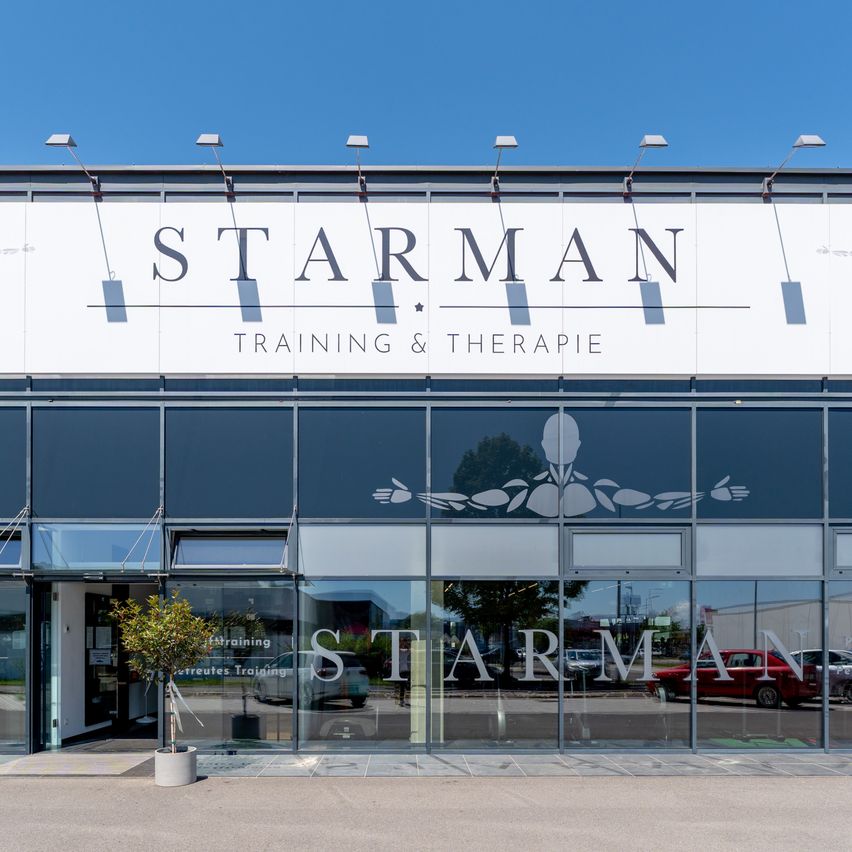 The front of Starman Training & Therapie, a modern building with glass walls and a white sign against a blue sky.