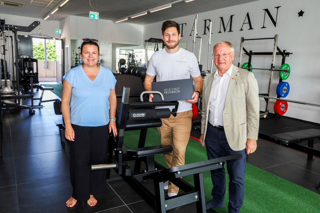 Three people stand in a gym, with a man holding a box labeled Neue Stadt Feldbach. The woman on the left wears a blue shirt and black pants, and the man on the right wears a beige jacket.