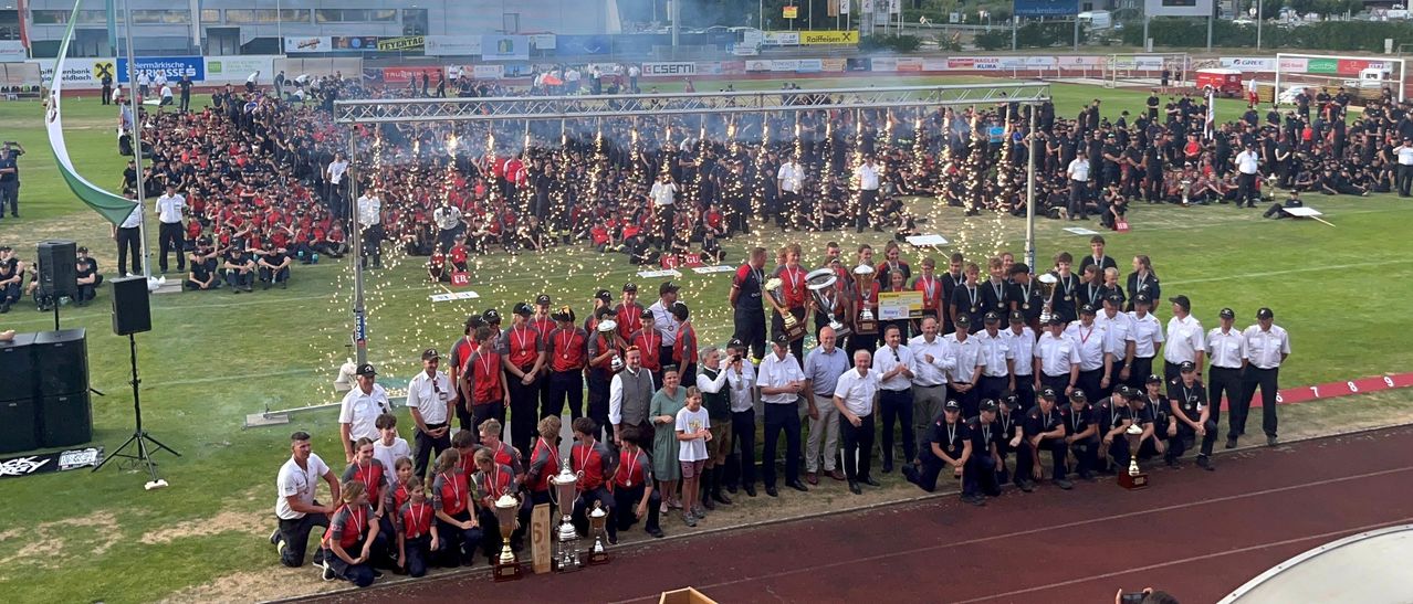 A large group of people, possibly a sports team, are celebrating on a field with trophies and confetti. Some are wearing red uniforms and medals, while others are in formal attire. The field is surrounded by a metal fence.