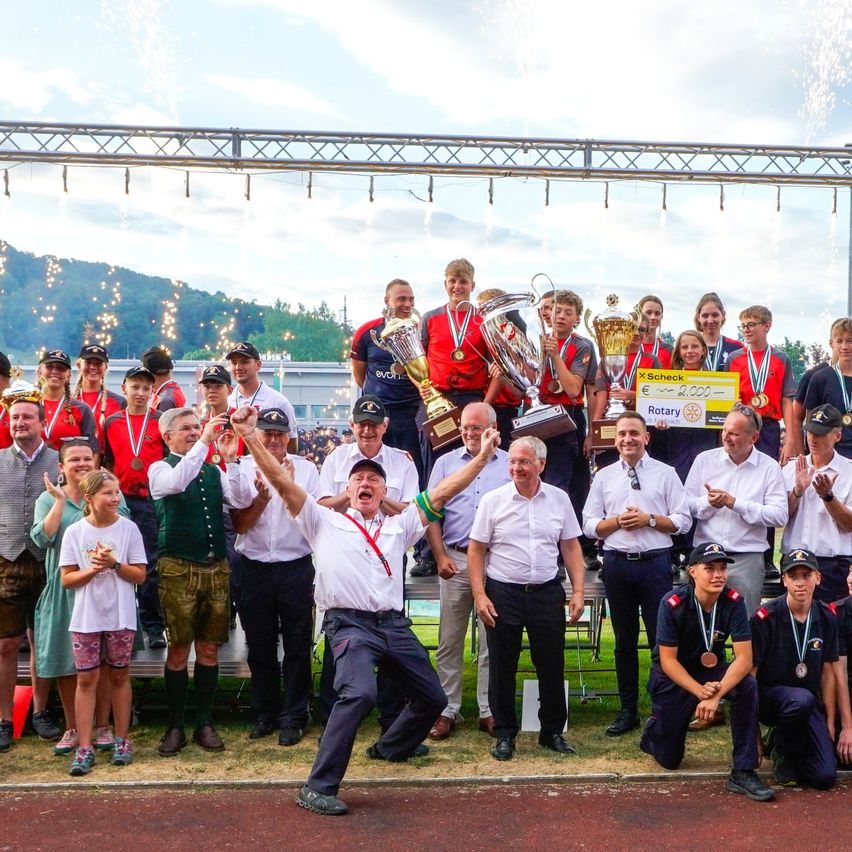A group of people in uniforms and medals are celebrating on a track with trophies and a large check in the background.