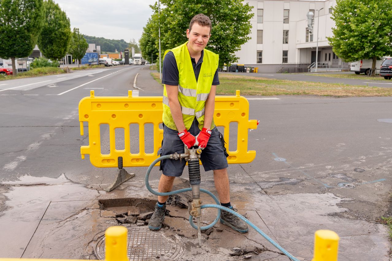 Ein Mann in einer gelben Weste und roten Handschuhen benutzt einen Presslufthammer, um einen Kanaldeckel auf einer Stadtstraße zu zerbrechen.