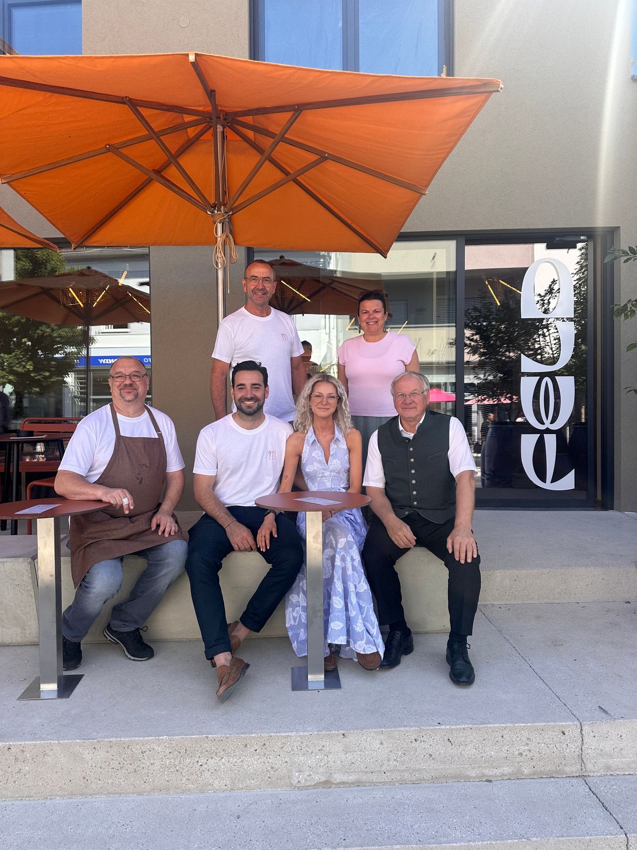 Fünf Personen sitzen vor einem Restaurant und lächeln für ein Foto. Ein Mann in einer Schürze steht links, während eine Frau in einem rosa T-Shirt hinter ihnen steht. Ein Regenschirm ist über ihnen.
