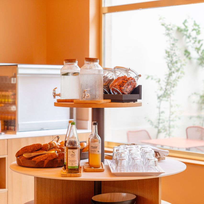 Ein hölzerner Tisch mit einem dreistufigen Ständer hält verschiedene Gegenstände, darunter Gläser mit Wasser, eine Flasche Saft, Gläser und eine Schüssel mit Brot. Im Hintergrund ist ein Fenster mit Blick auf eine Pflanze und Stühle zu sehen.