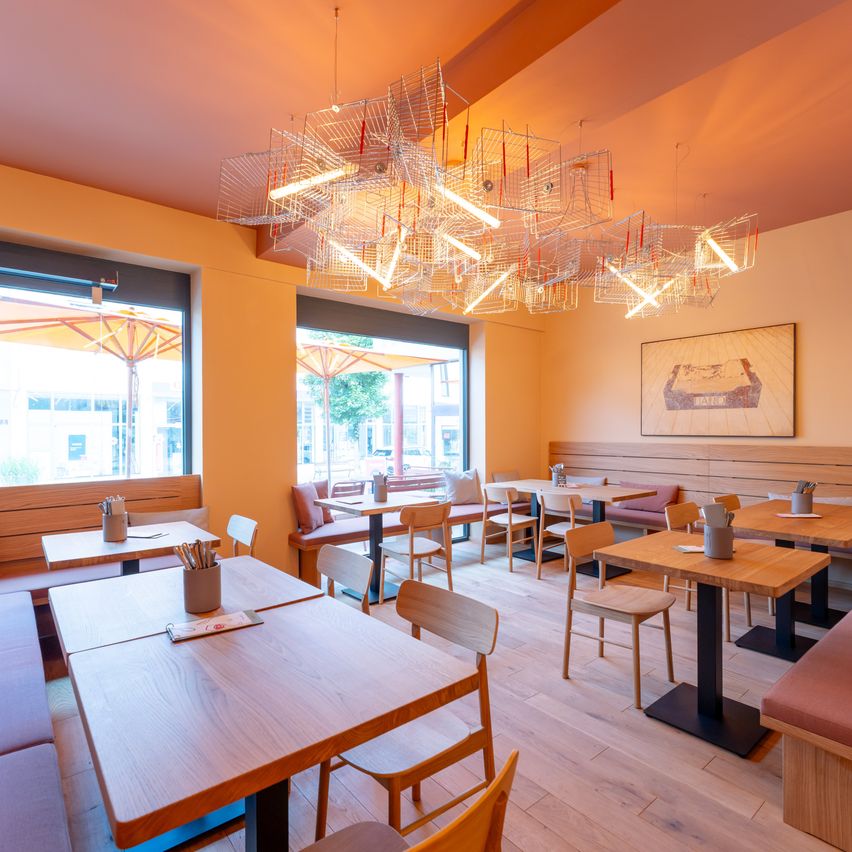 Ein Innenansicht eines modernen Restaurants mit Holztischen, Stühlen und einer einzigartigen Kronleuchter. Der Raum hat große Fenster mit Außenschirmen und einem Bilderrahmen an der Wand.