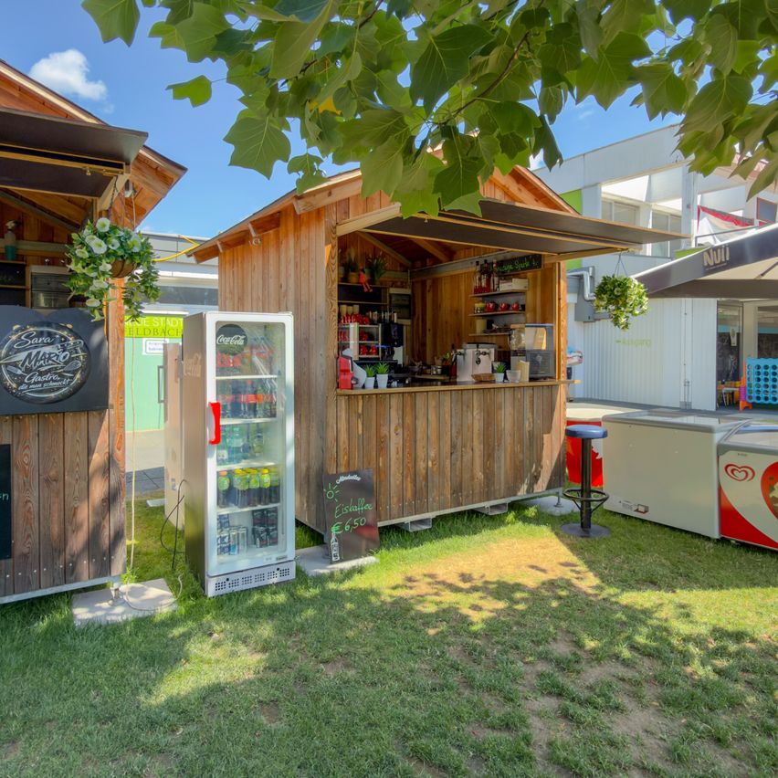 Ein kleiner hölzerner Kiosk mit einem Vordach und einem Kühlschrank voller Getränke. Er befindet sich auf einer Rasenfläche mit einem Gebäude im Hintergrund.