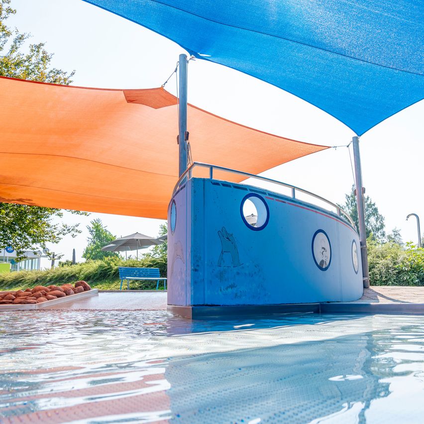 Ein blaues, schiffsförmiges Bauwerk mit einem runden Fenster schwimmt in einem Pool, bedeckt von blauen und orangefarbenen Sonnensegeln.